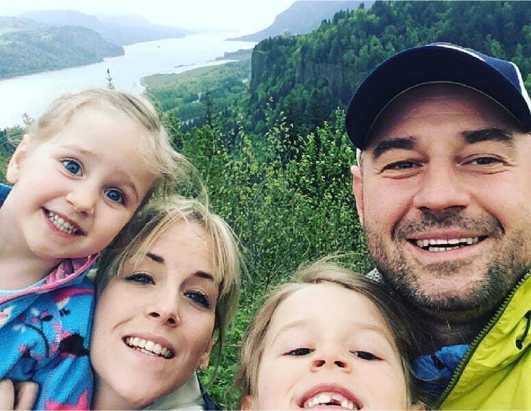Stacy Zelazny, her husband and their two young children, all pose for a selfie in front of a lush forested area that overlooks a distant. They are all smiling and dressed in hiking gear.